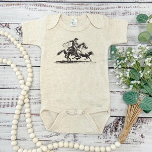 May include: A short-sleeved oatmeal-colored baby bodysuit featuring a black graphic print of a cowboy on horseback roping a calf. The garment has a round neckline and three metal snap closures at the bottom for easy access and secure fastening.