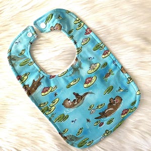 May include: A light blue baby bib with a pattern of otters swimming in a pond with lily pads. The bib has two white snaps on the back.