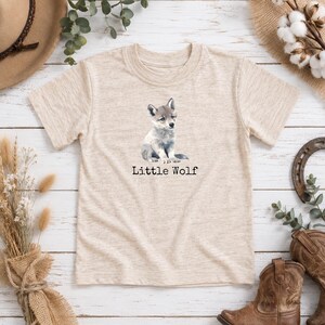 May include: A heathered beige short-sleeve t-shirt featuring a watercolour illustration of a grey wolf pup sitting in the centre. Below the illustration, the text Little Wolf is printed in a simple black serif font on the front of the shirt.