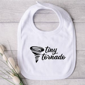 May include: White baby bib with a black graphic of a tornado and the words "tiny tornado". The bib has a rounded shape with a neck opening and a snap closure. The bib is made of soft fabric.