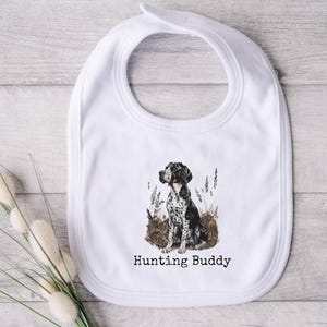 May include: White baby bib with a watercolor illustration of a black and white dog sitting in tall grass. The text "Hunting Buddy" is printed below the dog. The bib has a rounded shape and a snap closure.
