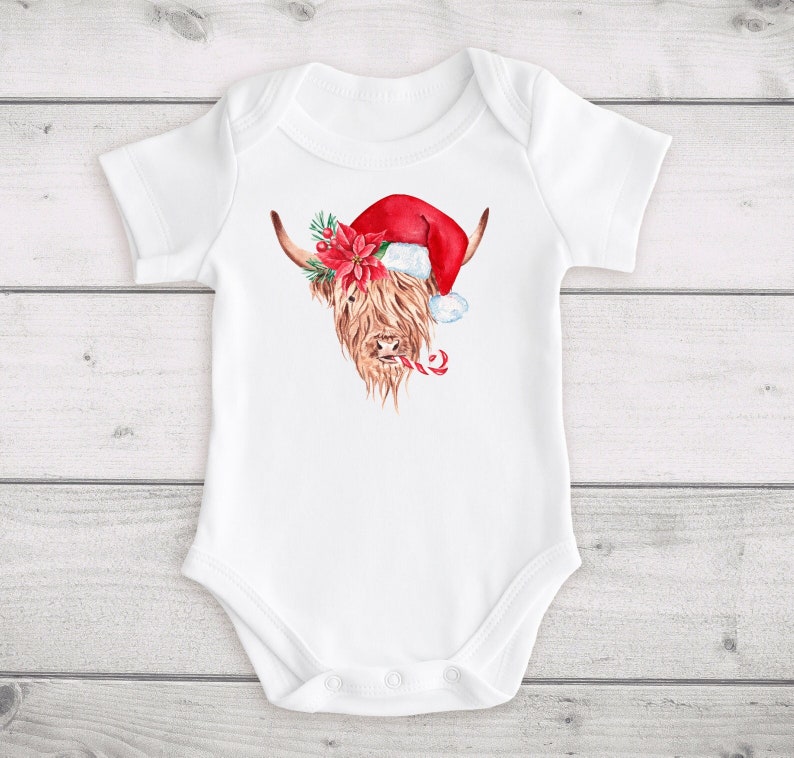 May include: White baby onesie featuring a watercolour illustration of a Highland cow wearing a Santa hat and holding a candy cane.