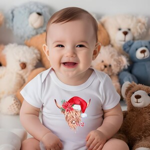 May include: A baby wearing a white onesie with a watercolour illustration of a Highland cow wearing a Santa hat and holding a candy cane. The baby is surrounded by plush toys.