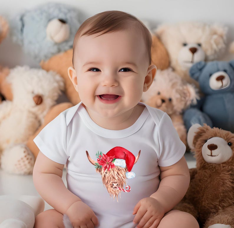 May include: A baby wearing a white onesie with a watercolour illustration of a Highland cow wearing a Santa hat and holding a candy cane. The baby is surrounded by plush toys.
