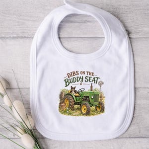 May include: A white baby bib featuring a graphic illustration of a green tractor with a dog sitting on the seat. The text above the tractor reads DIBS ON THE BUDDY SEAT in brown and green lettering. The design includes a windmill and grass.