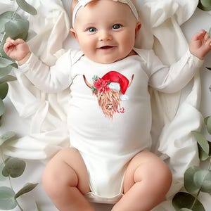 May include: A white baby onesie with a long sleeve design features a brown and white Highland cow wearing a red Santa hat. The cow is surrounded by red and green holly and a candy cane.