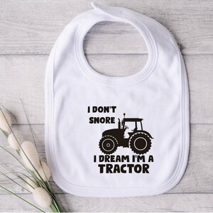 May include: White baby bib with a black tractor silhouette and the text "I don't snore I dream I'm a tractor".