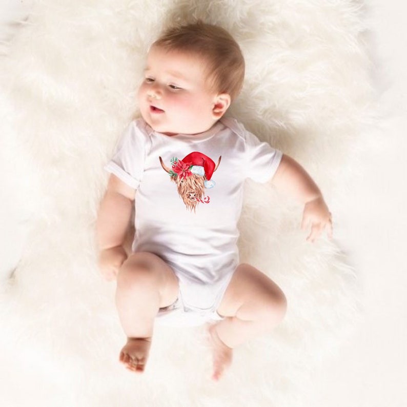 May include: A white baby onesie featuring a brown and white Highland cow wearing a red Santa hat with a green holly sprig and red berries. The onesie has short sleeves.