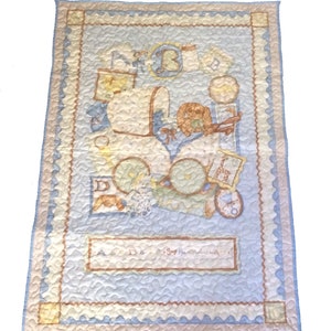 May include: A blue and cream baby quilt with a quilted pattern. The quilt features a baby carriage with a teddy bear and other baby items. The text "A B C - Say the alphabet with me" is printed on the quilt.