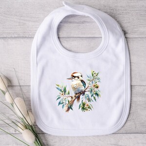 Peut inclure: Bavoir blanc pour bébé avec une illustration à l'aquarelle d'un oiseau kookaburra perché sur une branche avec des feuilles et des baies vertes. Le bavoir a une forme arrondie avec une ouverture pour le cou et une languette en haut.
