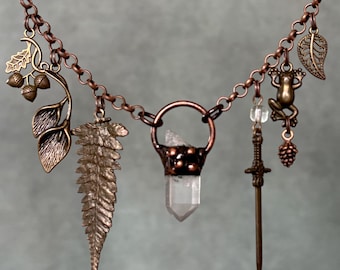 May include: A copper chain necklace featuring multiple charms including a clear quartz crystal point in a copper setting, a fern leaf, a sword, a frog, a pinecone, and a branch with acorns and leaves. The metal has a warm, metallic copper finish.