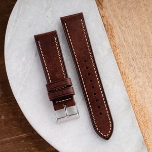 Pu&ograve; includere: Due cinturini per orologio in pelle marrone con cuciture bianche. I cinturini sono su una superficie di marmo bianco.