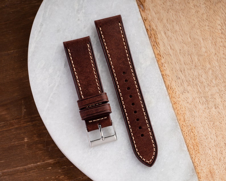 Pu&ograve; includere: Due cinturini per orologio in pelle marrone con cuciture bianche. I cinturini sono su una superficie di marmo bianco.