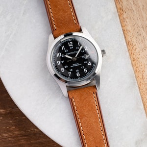 Pu&ograve; includere: Un orologio da polso argentato con quadrante nero e cinturino in pelle marrone. Il quadrante dell'orologio ha delle marcature bianche e il testo "Hamilton Khaki Automatic" stampato su di esso.