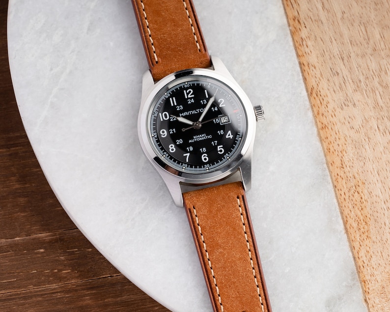 Pu&ograve; includere: Un orologio da polso argentato con quadrante nero e cinturino in pelle marrone. Il quadrante dell'orologio ha delle marcature bianche e il testo "Hamilton Khaki Automatic" stampato su di esso.