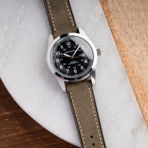 Pu&ograve; includere: Un orologio da polso argentato con quadrante nero e cinturino in pelle verde. Il quadrante dell'orologio ha delle marcature bianche e il testo "HAMILTON" e "KHAKI AUTOMATIC".