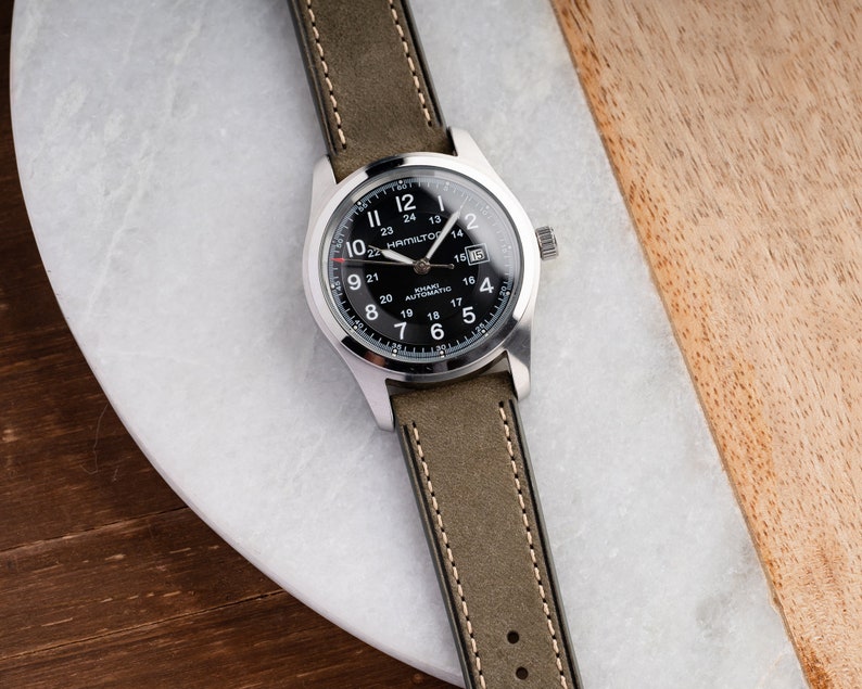 Pu&ograve; includere: Un orologio da polso argentato con quadrante nero e cinturino in pelle verde. Il quadrante dell'orologio ha delle marcature bianche e il testo "HAMILTON" e "KHAKI AUTOMATIC".