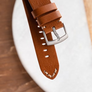 K&ouml;nnte beinhalten: Nahaufnahme eines braunen Leder-Uhrenarmbands mit silberner Schnalle. Das Armband hat sichtbare N&auml;hte und mehrere Verstelll&ouml;cher. Das Leder scheint hochwertig und langlebig zu sein, geeignet f&uuml;r eine Uhr.