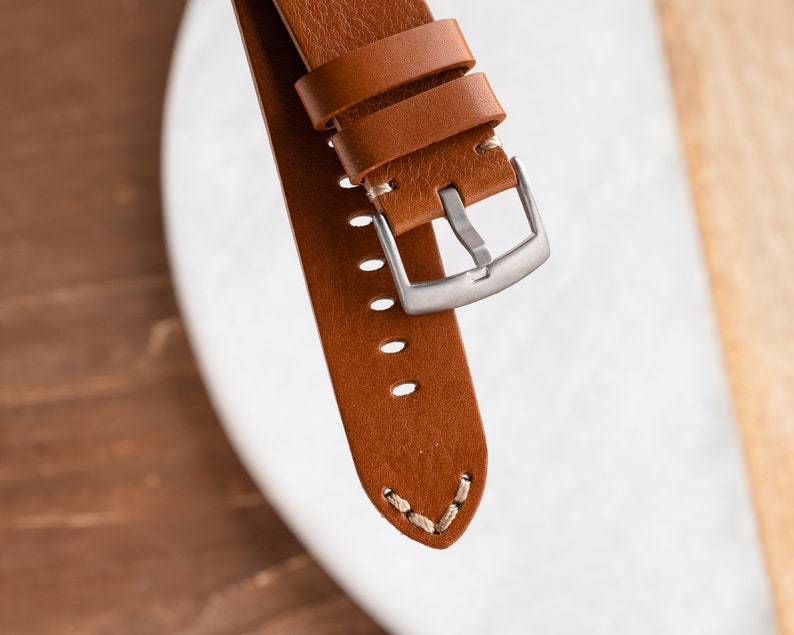 K&ouml;nnte beinhalten: Nahaufnahme eines braunen Leder-Uhrenarmbands mit silberner Schnalle. Das Armband hat sichtbare N&auml;hte und mehrere Verstelll&ouml;cher. Das Leder scheint hochwertig und langlebig zu sein, geeignet f&uuml;r eine Uhr.