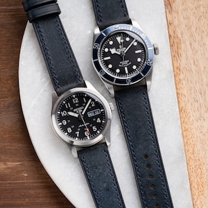 May include: Two wristwatches with black faces and blue leather bands. The watch on the left has a silver case and white markings. The watch on the right has a silver case with a black bezel and white markings. The watch on the right has the word "TUDOR" on the face.