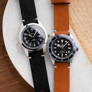 K&ouml;nnte beinhalten: Zwei Armbanduhren mit Lederarmb&auml;ndern. Eine hat ein schwarzes Zifferblatt mit wei&szlig;en Ziffern und dem Text "HAMILTON" und "KHAKI AUTOMATIC". Die andere Uhr hat ein schwarzes Zifferblatt mit einer blauen L&uuml;nette und dem Text "TUDOR".