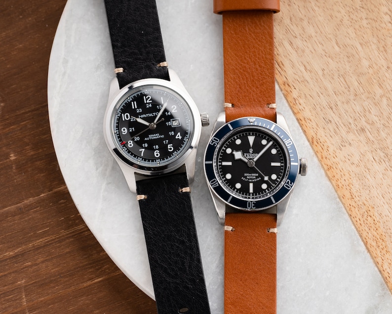 K&ouml;nnte beinhalten: Zwei Armbanduhren mit Lederarmb&auml;ndern. Eine hat ein schwarzes Zifferblatt mit wei&szlig;en Ziffern und dem Text "HAMILTON" und "KHAKI AUTOMATIC". Die andere Uhr hat ein schwarzes Zifferblatt mit einer blauen L&uuml;nette und dem Text "TUDOR".