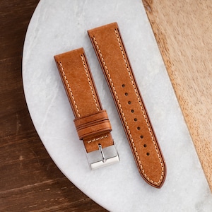 Pu&ograve; includere: Due cinturini per orologio in pelle marrone con cuciture bianche. I cinturini sono disposti su una superficie bianca.