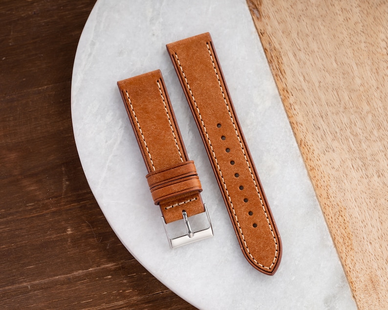 Pu&ograve; includere: Due cinturini per orologio in pelle marrone con cuciture bianche. I cinturini sono disposti su una superficie bianca.