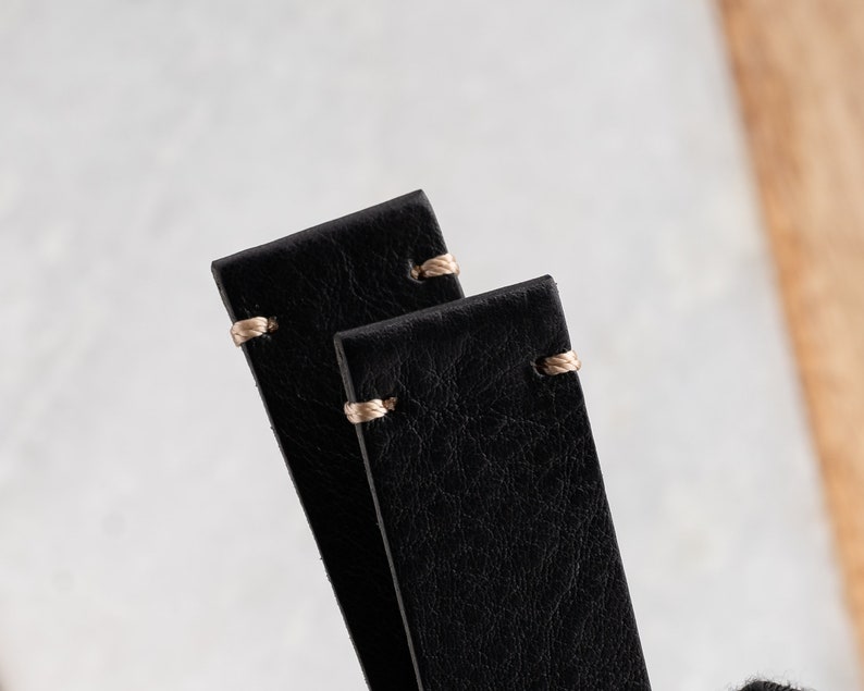K&ouml;nnte beinhalten: Nahaufnahme von zwei schwarzen Leder-Uhrenarmb&auml;ndern mit beigen N&auml;hten. Die Armb&auml;nder sind rechteckig mit abgerundeten Ecken und einer strukturierten Oberfl&auml;che. Die N&auml;hte sind oben an jedem Armband sichtbar und setzen einen Kontrast.