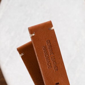 K&ouml;nnte beinhalten: Nahaufnahme von zwei braunen Leder-Uhrenarmb&auml;ndern mit wei&szlig;en N&auml;hten. Der Text "GENUINE LEATHER HANDMADE" ist auf einem Armband eingepr&auml;gt. Die Armb&auml;nder sind vor einem verschwommenen wei&szlig;en und h&ouml;lzernen Hintergrund platziert.