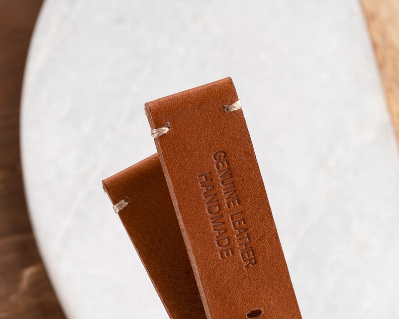 K&ouml;nnte beinhalten: Nahaufnahme von zwei braunen Leder-Uhrenarmb&auml;ndern mit wei&szlig;en N&auml;hten. Der Text "GENUINE LEATHER HANDMADE" ist auf einem Armband eingepr&auml;gt. Die Armb&auml;nder sind vor einem verschwommenen wei&szlig;en und h&ouml;lzernen Hintergrund platziert.