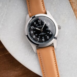 Może przedstawiać: Srebrny zegarek z czarną tarczą i brązowym sk&oacute;rzanym paskiem. Tarcza zegarka ma białe oznaczenia i napis "Hamilton Khaki Automatic" nadrukowany na niej.