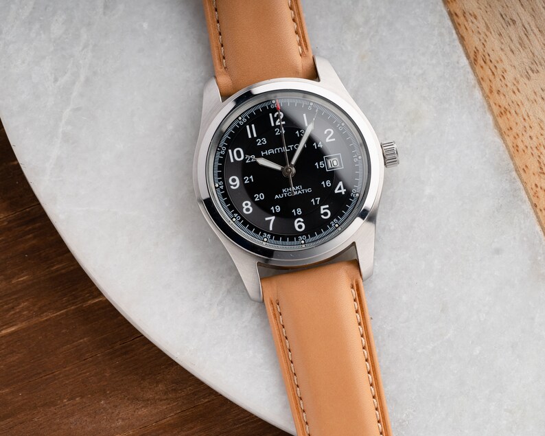 Może przedstawiać: Srebrny zegarek z czarną tarczą i brązowym sk&oacute;rzanym paskiem. Tarcza zegarka ma białe oznaczenia i napis "Hamilton Khaki Automatic" nadrukowany na niej.