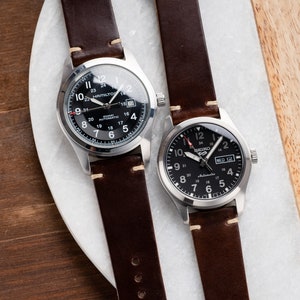 May include: Two wristwatches with black faces and brown leather bands. The watch on the left has a silver case and the text "Hamilton Khaki Automatic" on the face. The watch on the right has a silver case and the text "Seiko 5 Automatic" on the face.
