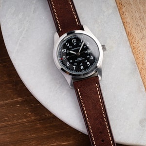 Pu&ograve; includere: Un orologio da polso argentato con quadrante nero e cinturino in pelle marrone. Il quadrante dell'orologio ha delle marcature bianche e il testo "Hamilton Khaki Automatic" stampato su di esso.