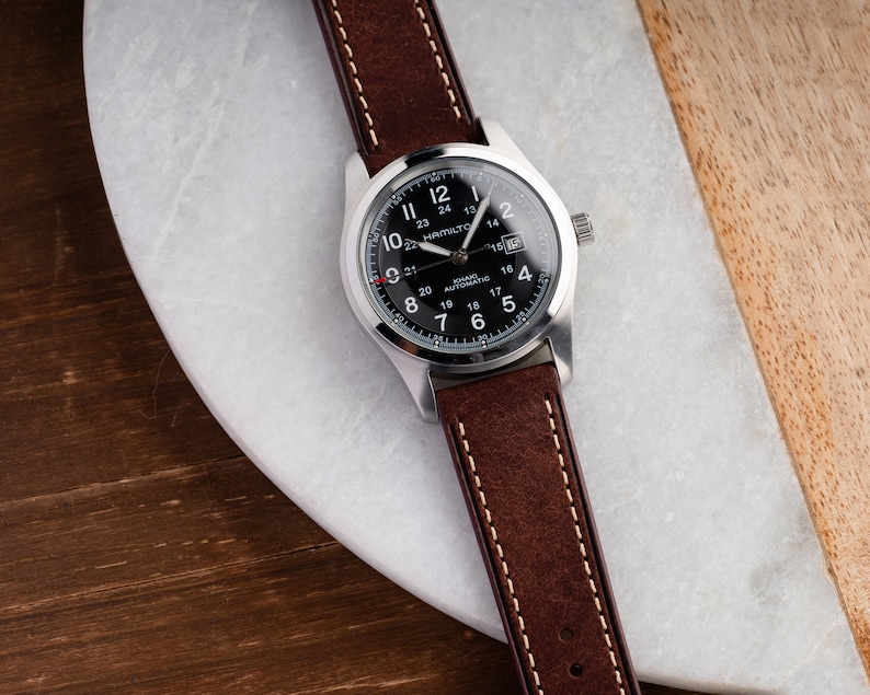 Pu&ograve; includere: Un orologio da polso argentato con quadrante nero e cinturino in pelle marrone. Il quadrante dell'orologio ha delle marcature bianche e il testo "Hamilton Khaki Automatic" stampato su di esso.