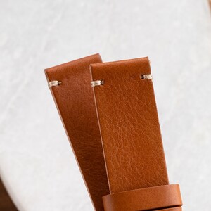 K&ouml;nnte beinhalten: Nahaufnahme von zwei braunen Lederarmb&auml;ndern. Die Armb&auml;nder sind in einem warmen, mittelbraunen Farbton gehalten und weisen eine sichtbare Maserung und Textur auf. Jedes Armband hat eine hellfarbige Naht am oberen Ende. Die Armb&auml;nder sind zum Betrachter hin abgewinkelt.