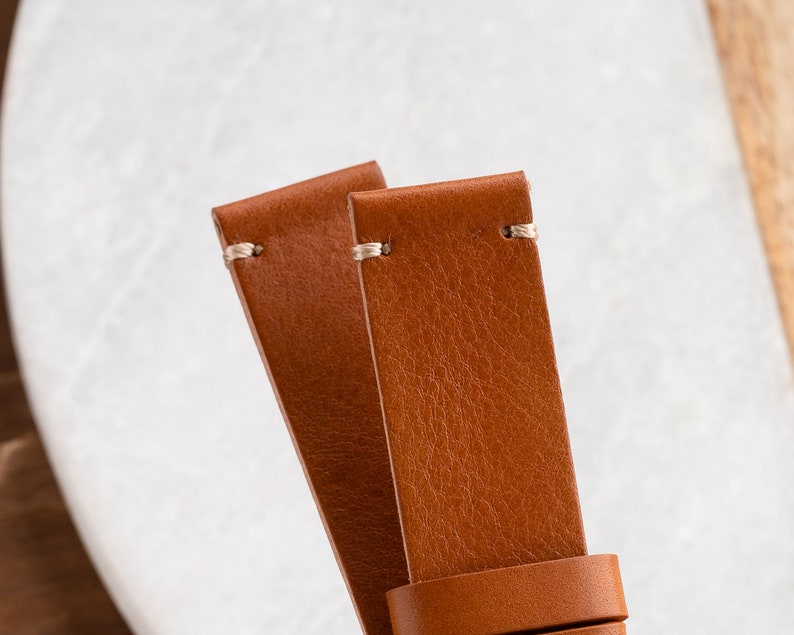 K&ouml;nnte beinhalten: Nahaufnahme von zwei braunen Lederarmb&auml;ndern. Die Armb&auml;nder sind in einem warmen, mittelbraunen Farbton gehalten und weisen eine sichtbare Maserung und Textur auf. Jedes Armband hat eine hellfarbige Naht am oberen Ende. Die Armb&auml;nder sind zum Betrachter hin abgewinkelt.
