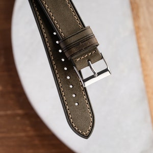 Pu&ograve; includere: Un cinturino per orologio in pelle verde con cuciture bianche e una fibbia argentata.