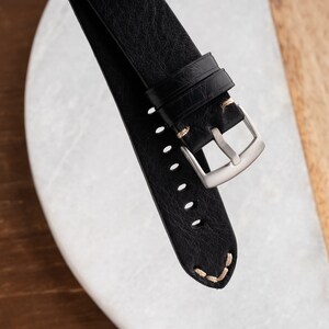 K&ouml;nnte beinhalten: Nahaufnahme eines schwarzen Leder-Uhrenarmbands mit silberner Schnalle. Das Armband hat sichtbare N&auml;hte in hellbeiger Farbe. Das Uhrenarmband liegt auf einer wei&szlig;en und braunen Oberfl&auml;che.