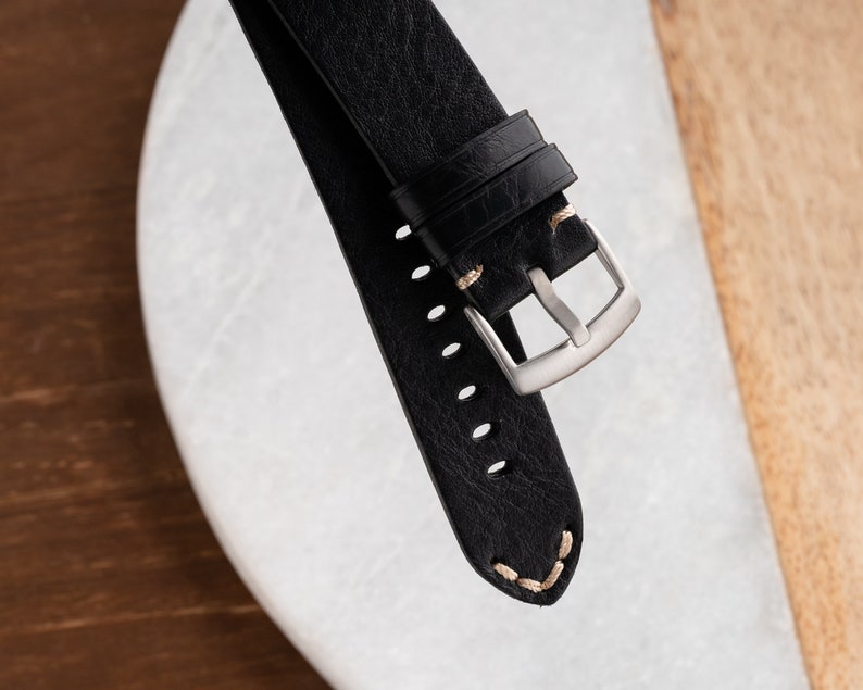 K&ouml;nnte beinhalten: Nahaufnahme eines schwarzen Leder-Uhrenarmbands mit silberner Schnalle. Das Armband hat sichtbare N&auml;hte in hellbeiger Farbe. Das Uhrenarmband liegt auf einer wei&szlig;en und braunen Oberfl&auml;che.