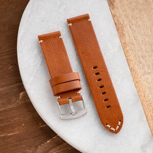 K&ouml;nnte beinhalten: Zwei braune Leder-Uhrenarmb&auml;nder mit wei&szlig;en N&auml;hten. Die Armb&auml;nder haben eine silberne Schnalle und werden auf einer wei&szlig;en und h&ouml;lzernen Oberfl&auml;che pr&auml;sentiert. Die Uhrenarmb&auml;nder sind neu.