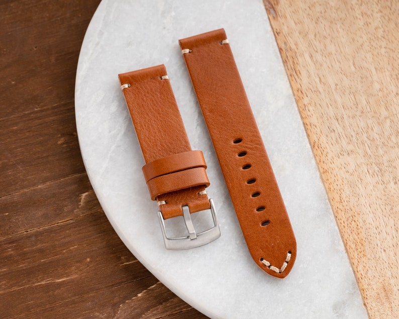 K&ouml;nnte beinhalten: Zwei braune Leder-Uhrenarmb&auml;nder mit wei&szlig;en N&auml;hten. Die Armb&auml;nder haben eine silberne Schnalle und werden auf einer wei&szlig;en und h&ouml;lzernen Oberfl&auml;che pr&auml;sentiert. Die Uhrenarmb&auml;nder sind neu.