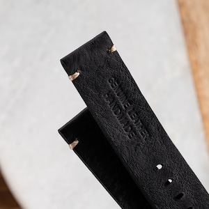 K&ouml;nnte beinhalten: Nahaufnahme von zwei schwarzen Lederarmb&auml;ndern. Die Armb&auml;nder sind mit hellfarbenem Faden gen&auml;ht und mit den Worten "GENUINE LEATHER HANDMADE" gepr&auml;gt. Das Leder hat ein strukturiertes Aussehen.