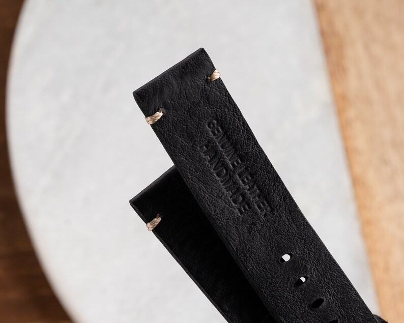K&ouml;nnte beinhalten: Nahaufnahme von zwei schwarzen Lederarmb&auml;ndern. Die Armb&auml;nder sind mit hellfarbenem Faden gen&auml;ht und mit den Worten "GENUINE LEATHER HANDMADE" gepr&auml;gt. Das Leder hat ein strukturiertes Aussehen.