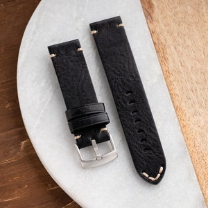 K&ouml;nnte beinhalten: Zwei schwarze Leder-Armbanduhren mit cremefarbenen N&auml;hten. Die Armb&auml;nder haben eine strukturierte Oberfl&auml;che und eine silberfarbene Schnalle. Eines der Armb&auml;nder hat eine Schlaufe. Sie liegen auf einer wei&szlig;en und holzmaserierten Oberfl&auml;che.