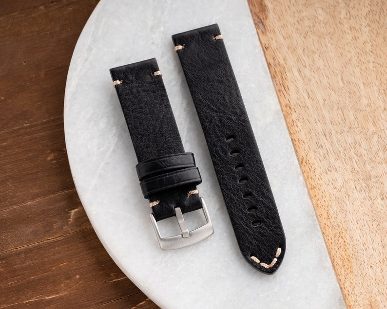 K&ouml;nnte beinhalten: Zwei schwarze Leder-Armbanduhren mit cremefarbenen N&auml;hten. Die Armb&auml;nder haben eine strukturierte Oberfl&auml;che und eine silberfarbene Schnalle. Eines der Armb&auml;nder hat eine Schlaufe. Sie liegen auf einer wei&szlig;en und holzmaserierten Oberfl&auml;che.