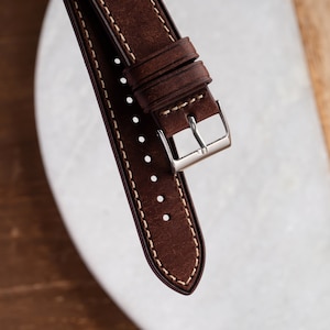 Pu&ograve; includere: Un cinturino da orologio in pelle marrone con cuciture bianche e una fibbia argentata.