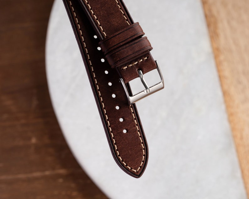 Pu&ograve; includere: Un cinturino da orologio in pelle marrone con cuciture bianche e una fibbia argentata.