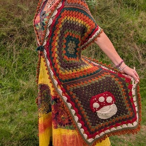 Può includere: Un cardigan all'uncinetto con un motivo a piastrelle granny multicolore nei toni del marrone, arancione, verde e rosso, con un evidente motivo a fungo bianco e rosso sul pannello posteriore, rifinito con un bordo smerlato color crema.